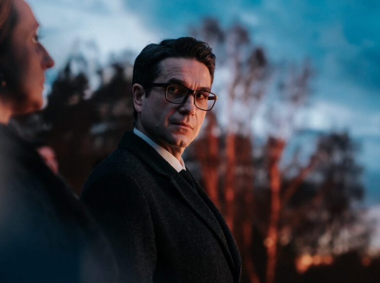 Kadr z filmu. Mężczyzna w ciemnym płaszczu i w okularach patrzy na kobietę, której fragment widać w rozmyciu. W tle nieostre drzewa i niebo.