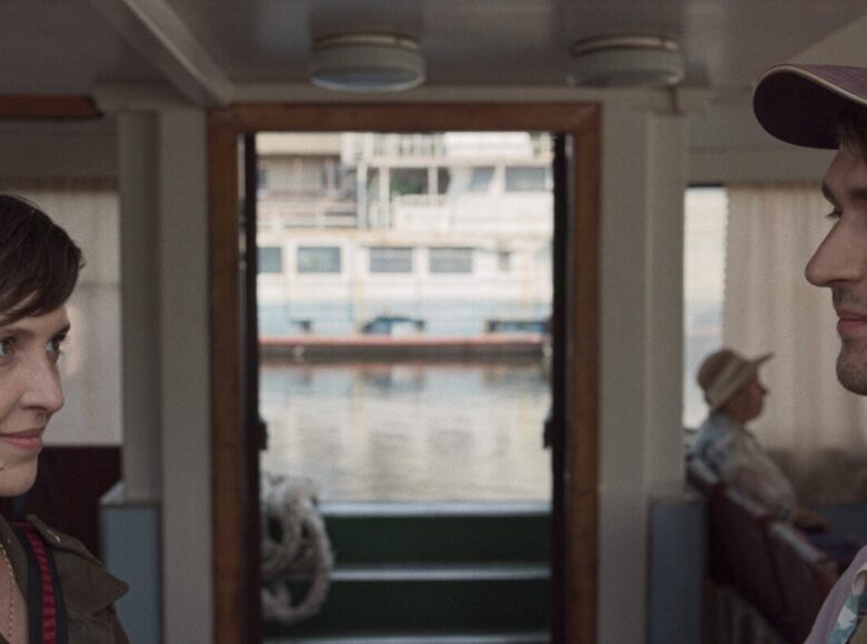 Pomieszczenie zamknięte. Na pierwszym planie zbliżenie na twarze kobiety i mężczyzny, którzy stoją naprzeciwko siebie. Za nimi w głębi pomieszczenia przy stole siedzi starsza kobieta. W tle okna wychodzące na zewnątrz.