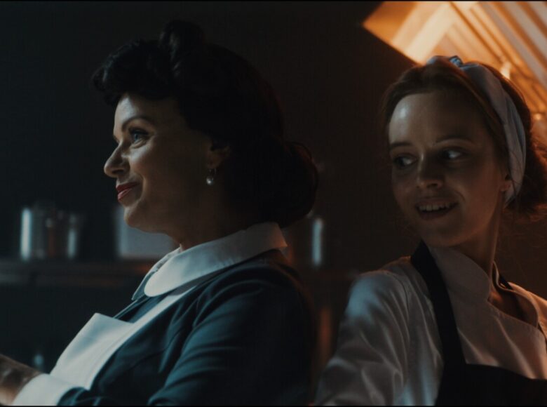 Wnętrze kuchni .na pierwszym planie oparte o siebie plecami dwie kobiety w fartuchach kucharskich. Wyglądają na zadowolone.