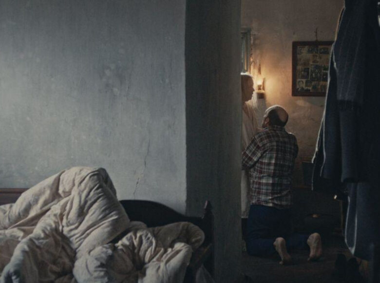 Ubogie pomieszczenie domowe. Na pierwszym planie łóżko ze skłębioną pościelą. W tle mężczyzna klęczący przed kobietą.