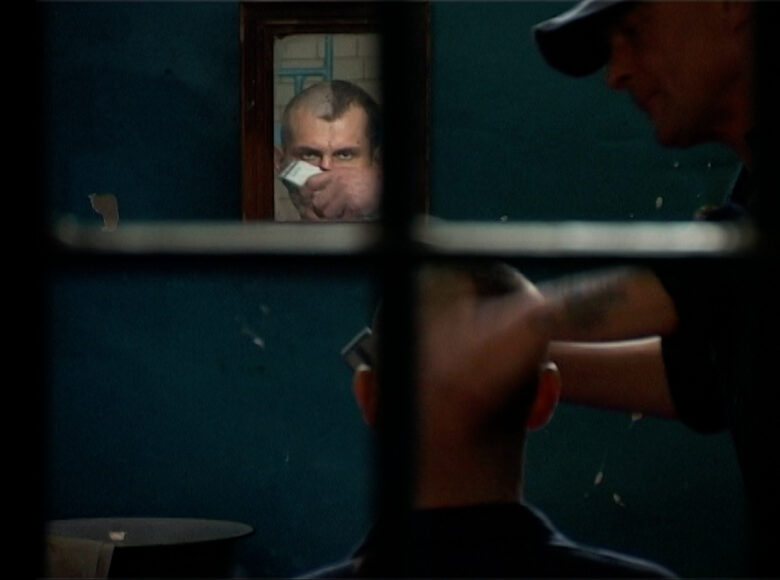 Cela więzienna. W niej dwóch mężczyzn. Trzeci mężczyzna zagląda do celi przez okienko.