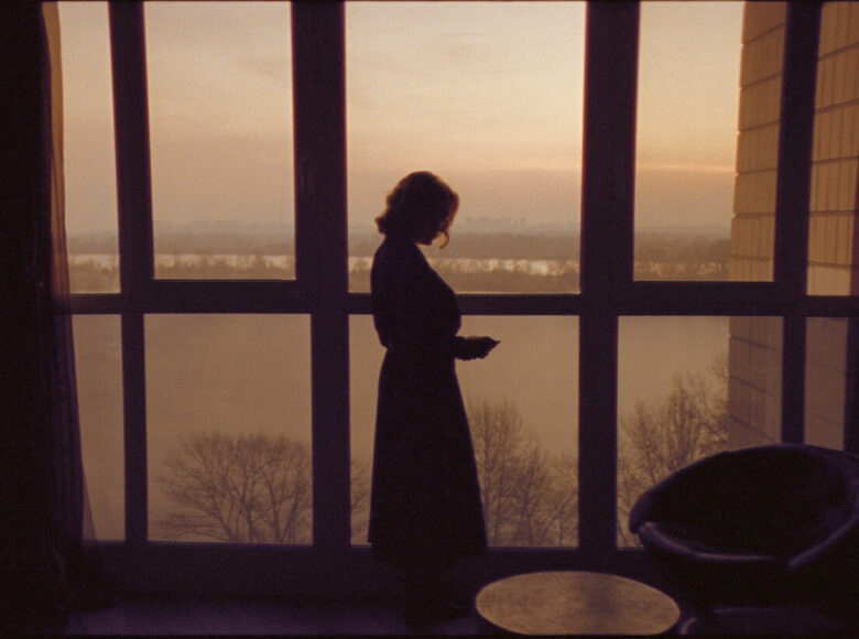 Wieczór, półmrok. Pomieszczenie domowe z wielkim oknem wychodzącym na drzewa i wodę. Przy oknie stoi kobieta.