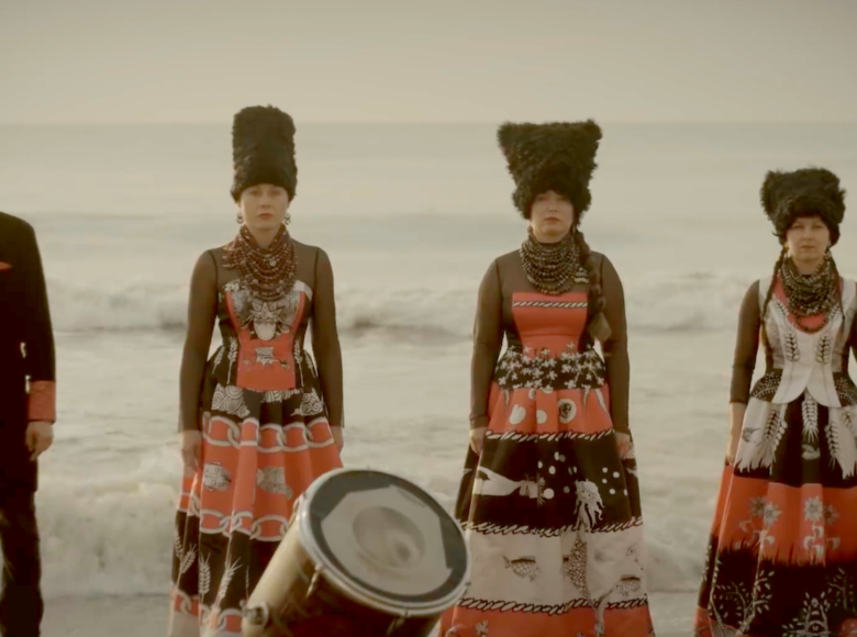 Na brzegu morza stoją frontem do obiektywu mężczyzna i trzy kobiety. Są ubrani w stroje etniczne. Jedna z kobiet trzyma w ręku wiolonczelę, przed drugą kobietą stoi bęben.