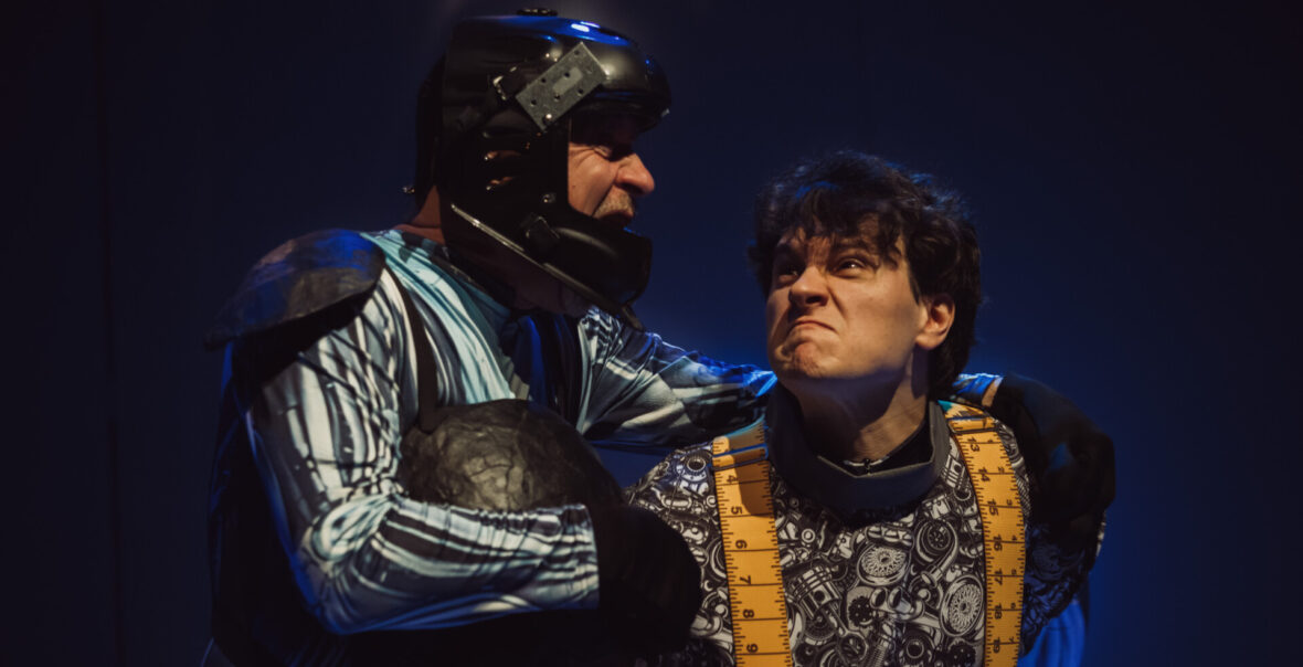 Spektakl teatralny. Zbliżenie na dwóch mężczyzn w kostiumach scenicznych. Za nimi ciemne tło.