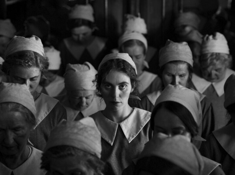 Na czarno białej fotografii grupa kobiet ubranych w takie same stroje robocze - fartuch i czepki. Pośrodku młoda kobieta ze zmęczoną twarzą, patrząca na wprost. Pozostałe kobiety mają spuszczone głowy i patrzą w dół.