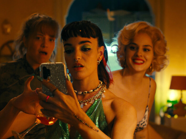 Trzy młode dziewczyny w imprezowych ubraniach patrzą na ekran telefonu komórkowego. Za nimi w tle wnętrze pokoju oświetlonego światłem lampek nocnych.