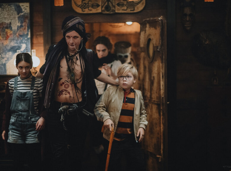 Kadr z filmu. Na pierwszym planie mężczyzna w fantazyjnym kostiumie prowadzi dziewczynkę i chłopca w codziennych ubraniach. Za nimi pomieszczenie z drewnianymi ścianami, na ścianach mapa oraz etniczne maski i obrazy.