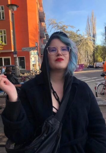 Jasny, wiosenny dzień w mieście. Na pierwszym planie młoda dziewczyna z niebieskimi włosami, w okularach i czarnej kurtce. W dłoni trzyma papierosa.