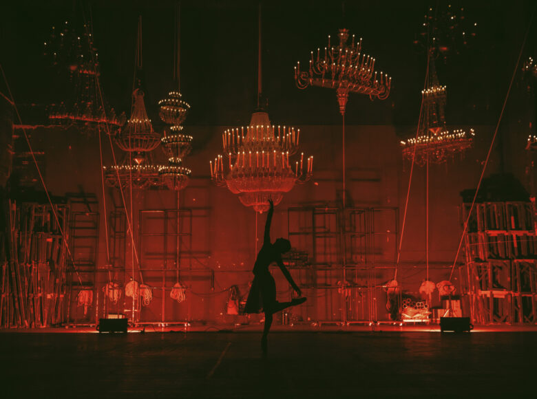 W pomieszczeniu tancerka w baletowej pozie. Dookoła wiszą ozdobne żyrandole.