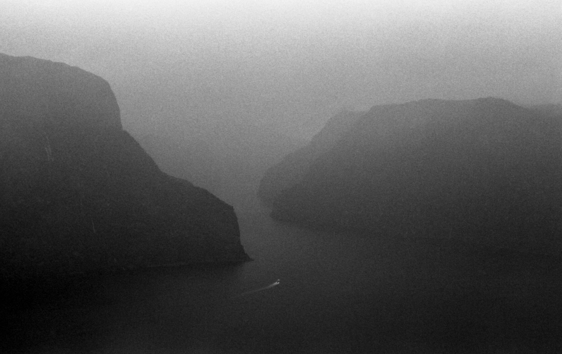 Na czarno-białej fotografii widzimy spowity we mgle fiord, otoczony wysokimi górami. Do&nbsp;przesmyka między tymi górami, zbliża się widoczna w&nbsp;oddali mała motorówka, zostawiając za&nbsp;sobą w&nbsp;wodzie jasny ślad, załamujących się fal.