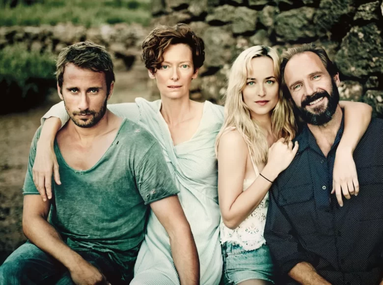 Na ławce siedzą dwie kobiety i dwaj mężczyźni w letnich ubraniach. Za nimi trawa, krzaki i fragment muru.