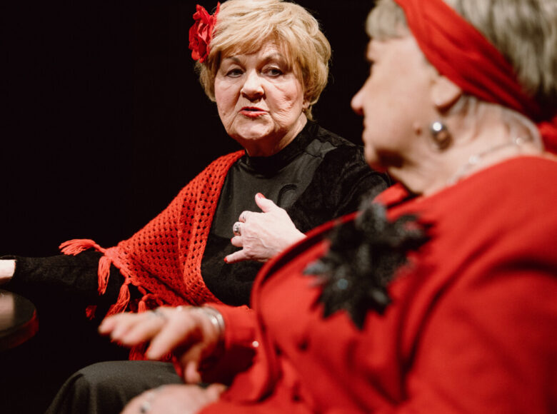 Zbliżenie na dwie kobiety siedzące obok siebie przy stole. Za nimi czarne tło.