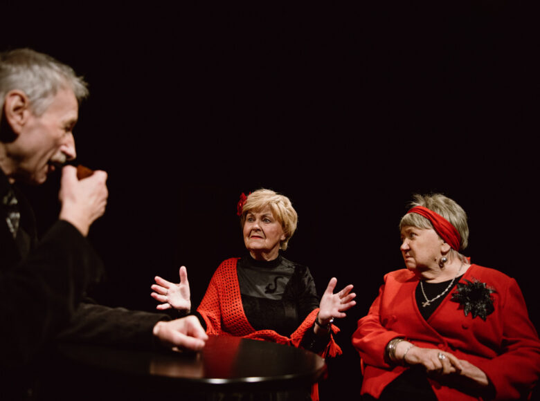 Dwie kobiety siedzą przy stole. Naprzeciwko nich siedzi mężczyzna z fajką w dłoni.