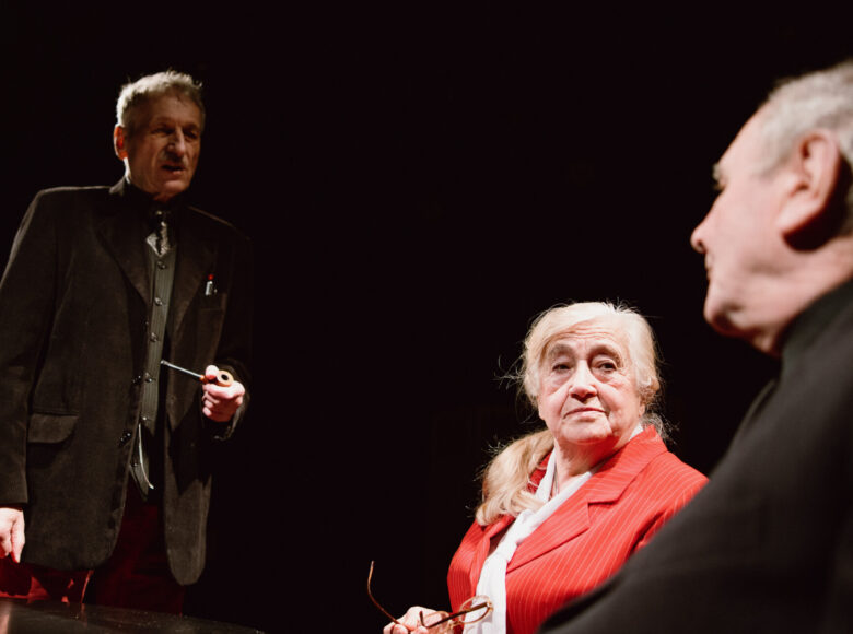 Mężczyzna i kobieta siedzą przy stole. Naprzeciwko nich stoi drugi mężczyzna z fajką w dłoni.