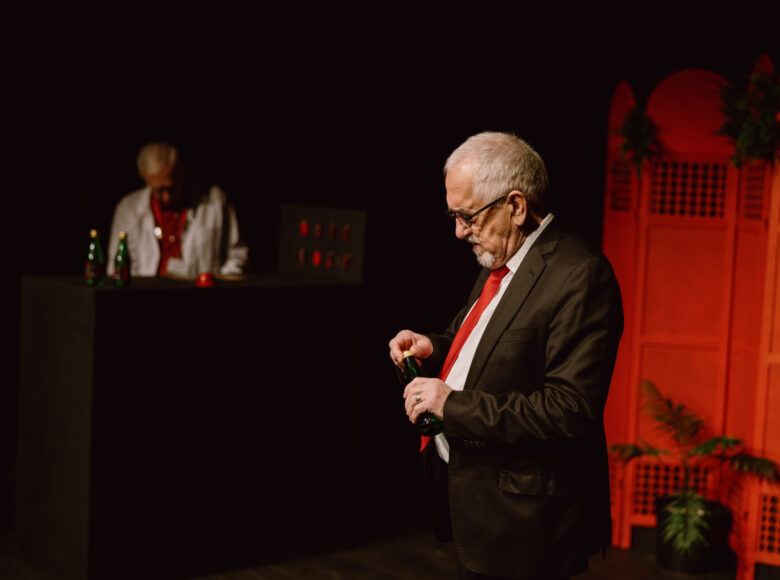 W ciemnym pomieszczeniu stoi starszy mężczyzna w garniturze, w dłoniach trzyma butelkę z jakimś napojem. Za nim w głębi czarny bar na tle czarnej ściany. Za barem stoi drugi mężczyzna. Obok baru stoi czerwony parawan.