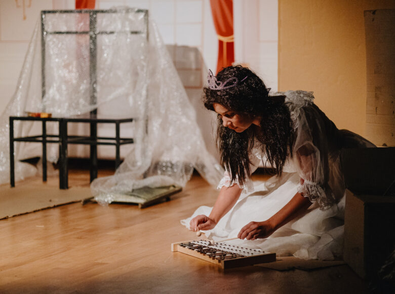 Spektakl teatralny. Na scenie kobieta w bajkowym kostiumie gra na cymbałkach. Za nią minimalistyczna dekoracja z tektury i folii bąbelkowej.