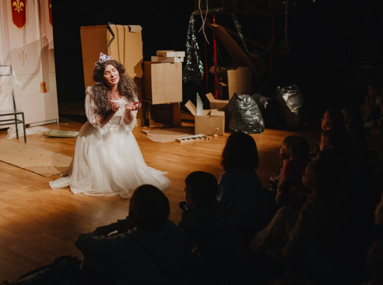 Spektakl teatralny. Na scenie kobieta w bajkowym kostiumie księżniczki. Za nią minimalistyczna dekoracja z pudeł tekturowych.