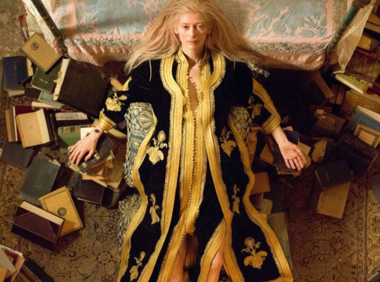 Kobieta w luksusowym szlafroku leży na dywanie, wokół niej porozrzucane książki.