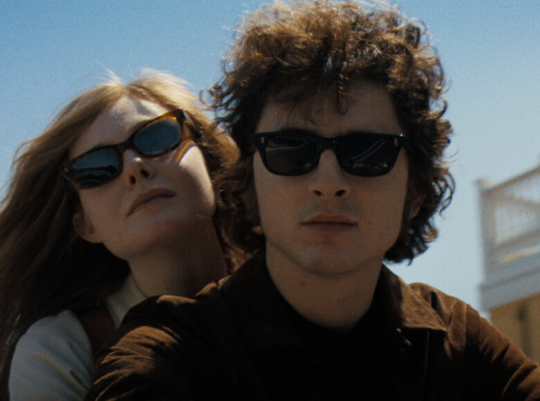 Na pierwszym planie twarz chłopaka. Za nim twarz obejmującej go od tyłu dziewczyny. Oboje w okularach słonecznych. Za nimi jasne niebo i fragment budynku.
