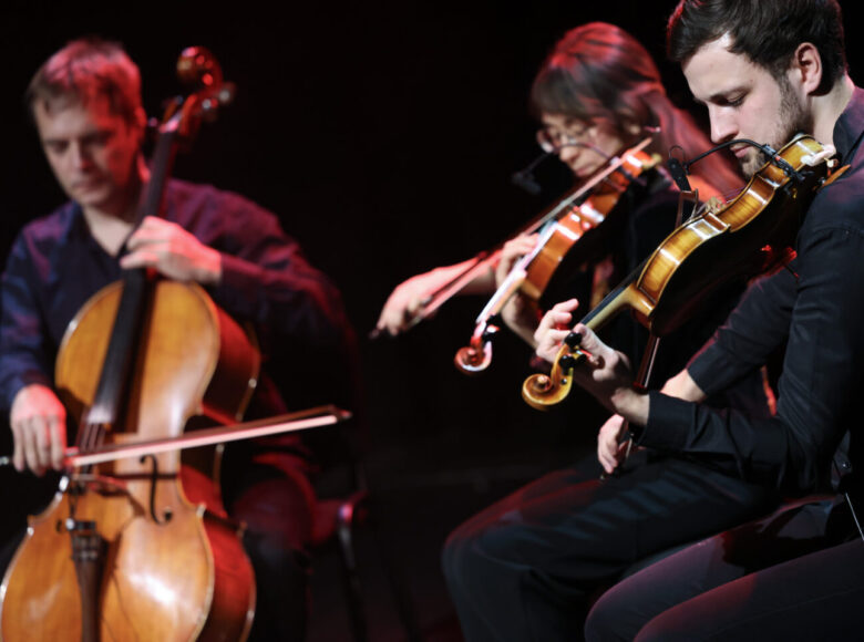 Na scenie na czarnym tle troje muzyków. Kobieta i mężczyzna grający na skrzypcach oraz drugi z mężczyzn grający na wiolonczeli.