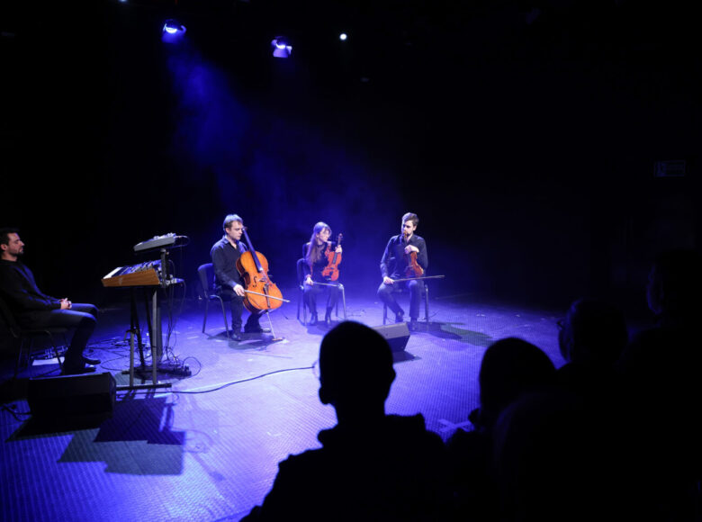 Na scenie siedzi czworo muzyków z instrumentami - kobieta i jeden z mężczyzn trzymają skrzypce, drugi mężczyzna trzyma wiolonczelę, trzeci mężczyzna siedzi przy syntezatorze dźwięku.