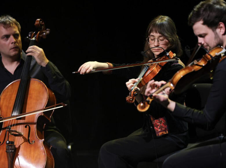 Na scenie na czarnym tle troje muzyków. Kobieta i mężczyzna grający na skrzypcach oraz drugi z mężczyzn grający na wiolonczeli.