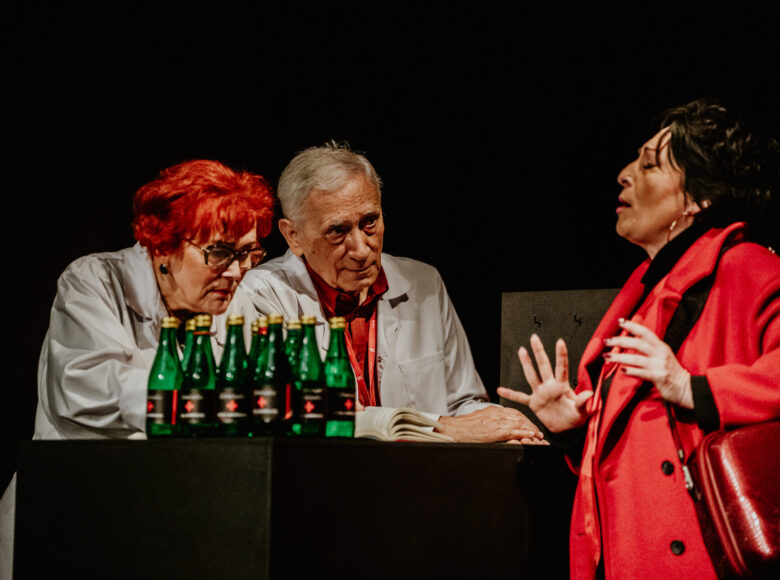 Ciemne pomieszczenie z punktowym światłe. Przy barze stoją dwie starsze kobiety i starszy mężczyzna, wszyscy elegancko ubrani. Na barze stoi rząd szklanych butelek z wodą mineralną.