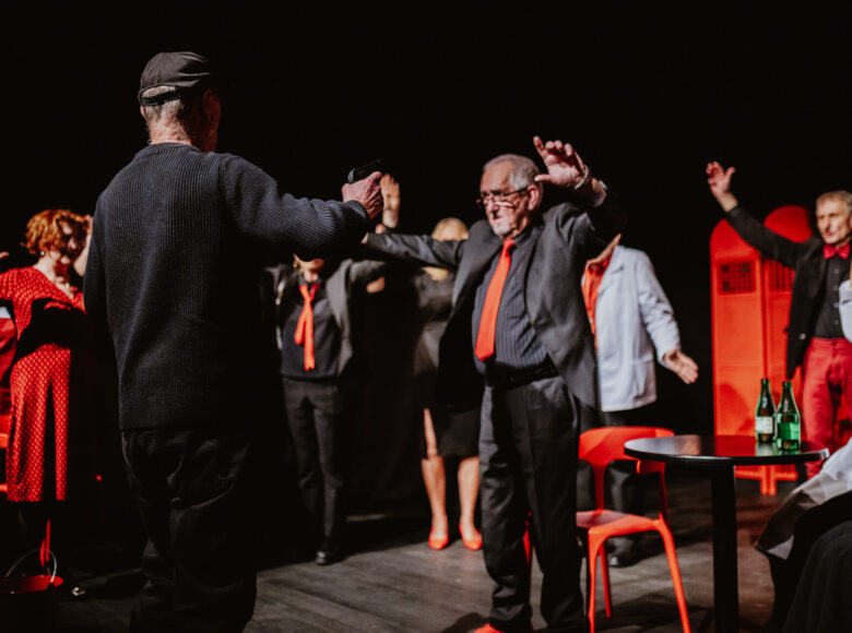 Ciemne pomieszczenie z punktowym oświetleniem. Grupa starszych elegancko ubranych osób stoi w kręgu. Unoszą ręce do góry.