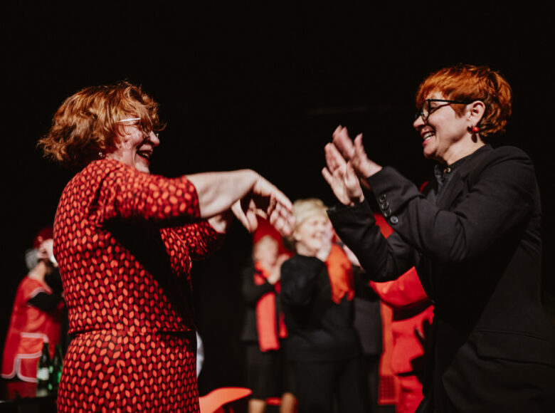 Ciemne pomieszczenie z punktowym światłem. Dwie roześmiane starsze kobiety stoją naprzeciwko siebie i wyciągają do siebie ręce. W tle grupa innych starszych kobiet.