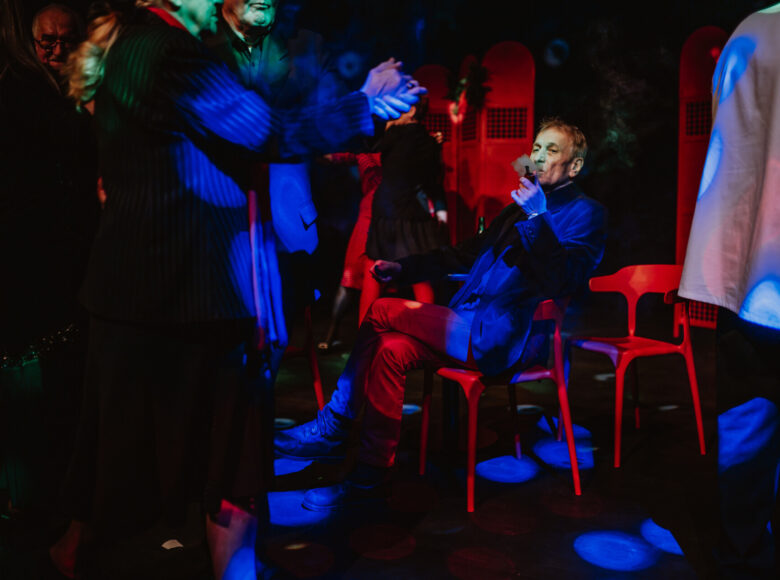 Ciemne pomieszczenie punktami niebieskiego światła. Kilka osób tańczy, obok mężczyzna siedzi na krześle i pali fajkę.