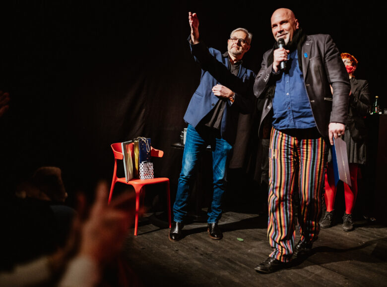 Na scenie mężczyzna przemawia do mikrofonu. Obok stoi drugi mężczyzna wskazując na coś wyciągniętą dłonią. Za nimi kobieta z czerwoną różą w dłoni. Jest ciemno, oświetlenie jest punktowe.