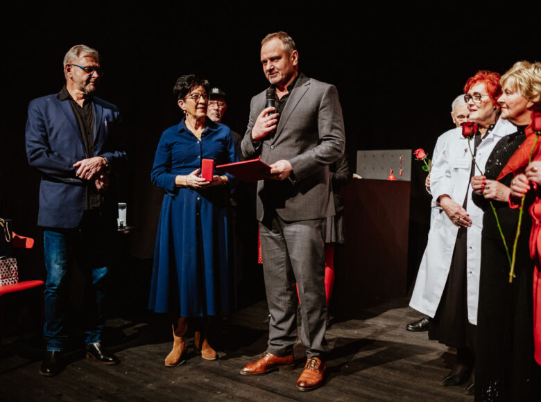 Na scenie stoi grupa elegancko ubranych starszych kobiet i mężczyzn. Jeden z mężczyzn przemawia do mikrofonu. Kilka kobiet trzyma w dłoniach czerwone róże.