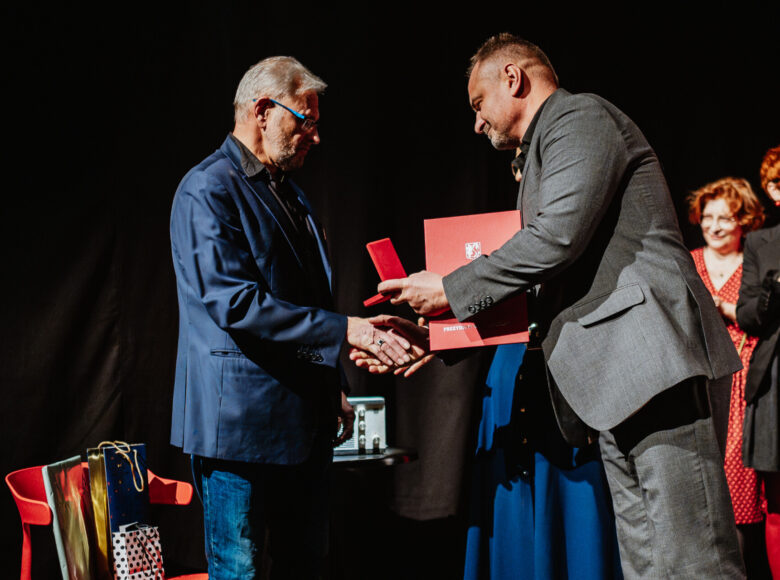 Na scenie dwóch elegancko ubranych mężczyzn mężczyzn. Jeden ściska drugiemu dłoń w geście gratulacji, a drugą ręką podaje mu pudełko z medalem i dyplom. Za nimi w tle stoją trzy elegancko ubrane kobiety.