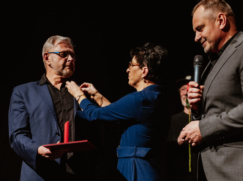 Na scenie stoi kobieta w eleganckiej sukience i dwóch mężczyzn w garniturach. Kobieta przypina jednemu z mężczyzn odznaczenie do klapy marynarki.