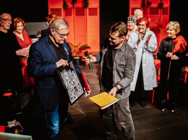 Ciemne wnętrze z punktowym oświetleniem. Mężczyzna pokazuje trzymany w rękach obrazek drugiemu mężczyźnie z mikrofonem. Obu mężczyznom przygląda się grupa starszych osób.