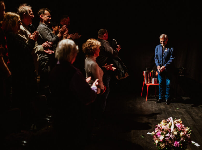 Sala teatralna. Publiczność oklaskuje na stojąco kłaniającego się mężczyznę w niebieskim garniturze. Przed nim na podłodze stoi duży bukiet kwiatów. Sala jest ciemna z punktowym oświetleniem.