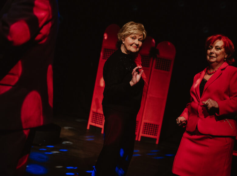 Ciemne pomieszczenie z punktowym światłem. Dwie tańczące starsze kobiety, elegancko ubrane.