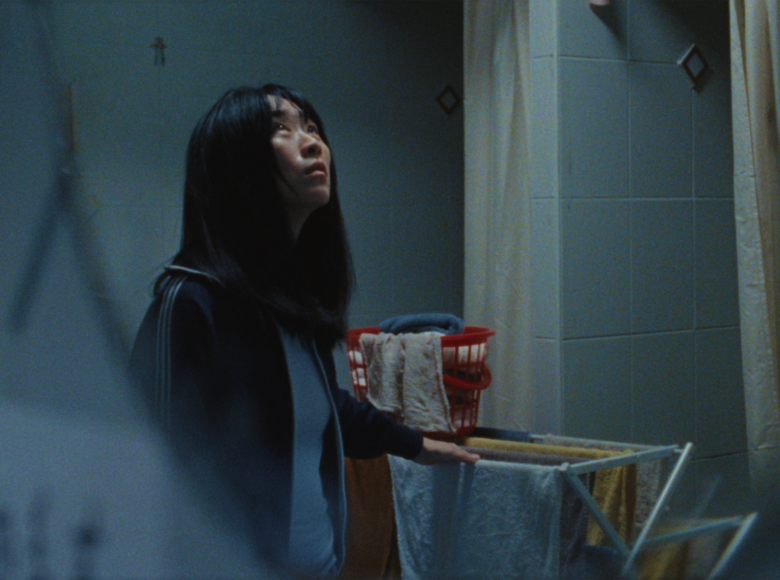 Łazienka. Obok suszarki z suszącym się praniem stoi dziewczyna o azjatyckich rysach twarzy ubrana w bluzę dresową. Dziewczyna patrzy w górę.