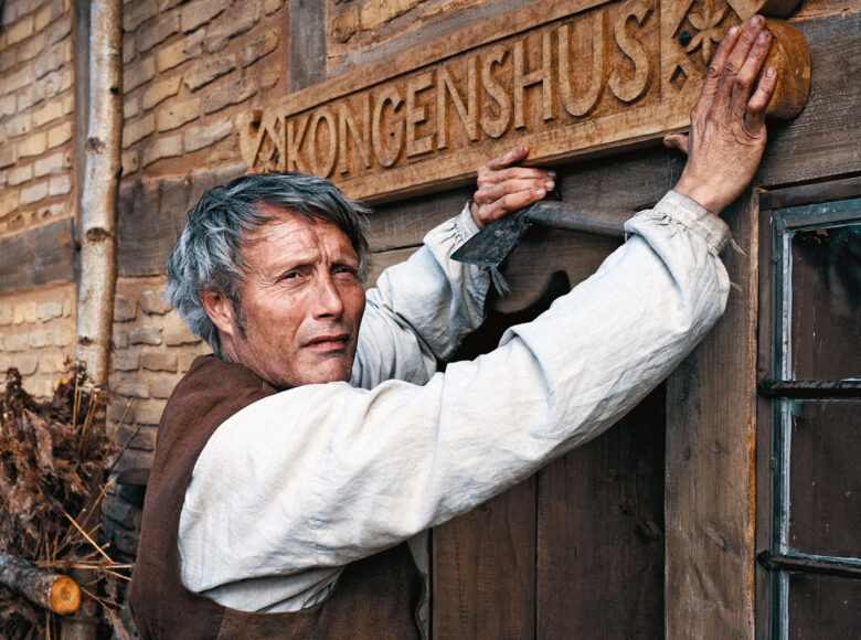 Przed wiejską, drewnianą chatą stoi mężczyzna w średnim wieku ubrany w starodawną chłopską koszulę.