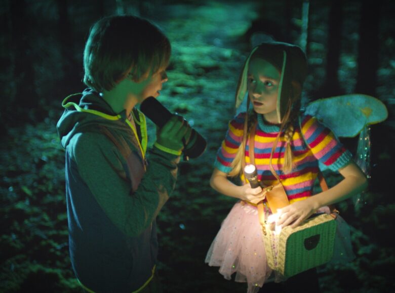 Las lub park, jest ciemno. Na pierwszym planie dziewczynka i chłopiec stoją naprzeciwko siebie. Chłopiec ma bluzę z kapturem. Dziewczynka ma kolorowy sweterek i różową spódniczkę z tiulu, czapkę z długimi zwierzęcymi uszami oraz skrzydełka przyczepione do ramion. Oboje mają w rękach latarki. Dziewczynka ma także wiklinowy kuferek, przewieszony na pasku przez ramię.