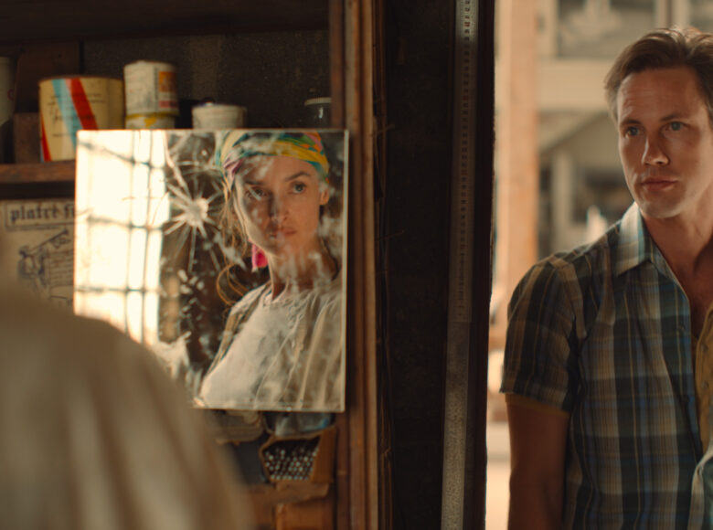 Młody chłopak w letniej koszuli stoi w drzwiach z ulicy do pomieszczenia. Obok drzwi półka na której stoją puszki z farbą. Na półce wisi także lustro, w którym odbija się twarz młodej dziewczyny.