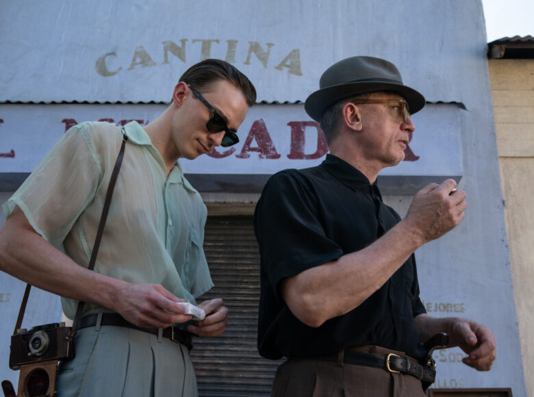 Dwaj mężczyźni, jeden dojrzały drugi młody, stoją obok siebie. Ubrani są w letnie ubrania. Za nimi widać kamienicę z witryną sklepu zasłoniętą żaluzją.