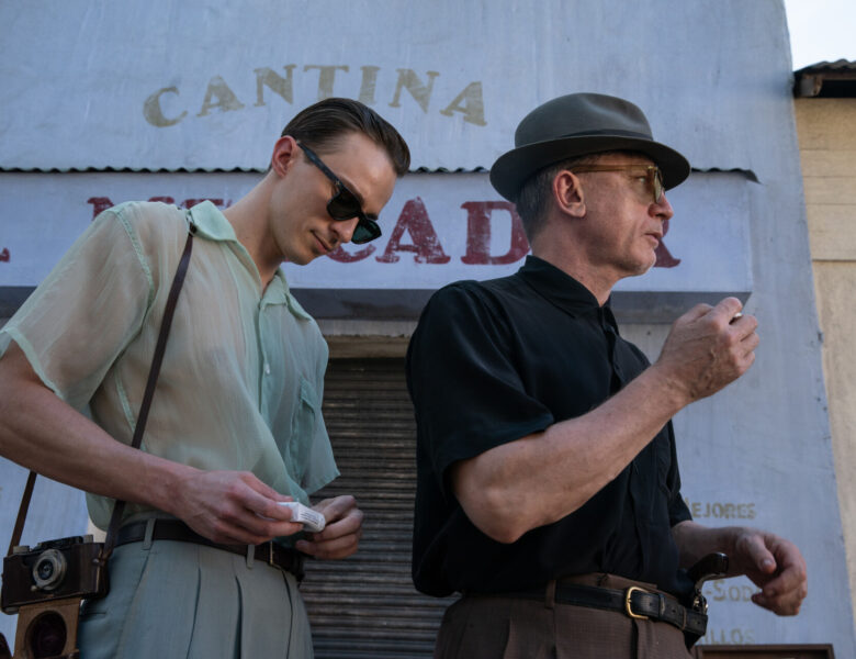 Dwaj mężczyźni, jeden dojrzały drugi młody, stoją obok siebie. Ubrani są w letnie ubrania. Za nimi widać kamienicę z witryną sklepu zasłoniętą żaluzją.