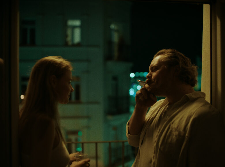 Jest wieczór. Kobieta i mężczyzna stoją w pokoju na tle otwartego okna. Mężczyzna pali papierosa. Za oknem widać kamienice, światła w oknach i czarne niebo.