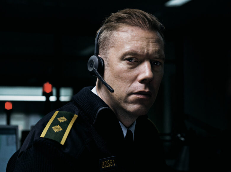 Zbliżenie na twarz i ramiona mężczyzny w mundurze, ze słuchawkami na uszach. W tle ciemność, z której wyłaniają się fragmenty niedookreślonych obiektów. Gdzieniegdzie kilka punktów czerwonego światła.