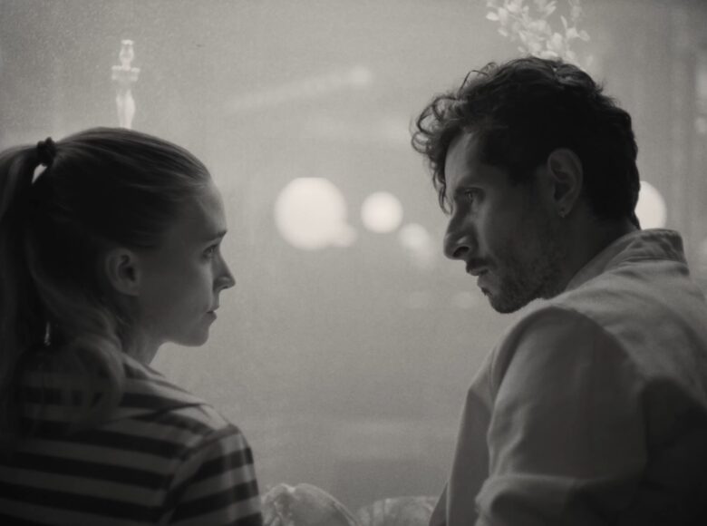 Na pierwszym planie młoda dziewczyna i młody mężczyzna stoją razem patrząc na siebie. Za nimi niedookreślone tło na którym można dostrzec kilka punktów światła.