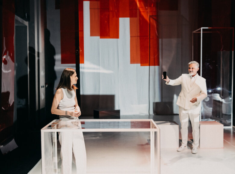 Spektakl teatralny. Na scenie kobieta i mężczyzna ubrani na biało. Stoją w pewnej odległości od siebie. Mężczyzna wyciąga w kierunku kobiety rękę z telefonem komórkowym.