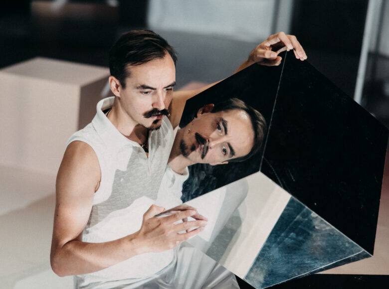 Spektakl teatralny. Na scenie siedzi mężczyzna trzymając w rękach duży sześcian zbudowany z luster. Twarz mężczyzny odbija się w sześcianie.