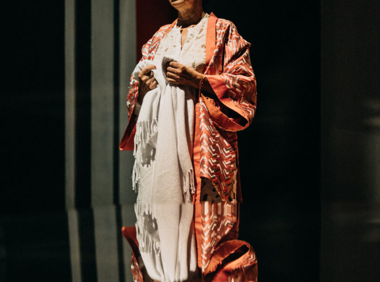 Spektakl teatralny. Starsza kobieta stoi na ciemnym tle. Przed nią leży lustro, w którym widzimy także jej odbicie.