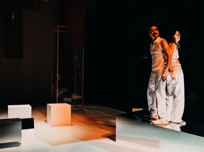 Spektakl teatralny. Kobieta i mężczyzna oświetleni punktowo stoją oparci o siebie plecami. Oboje ubrani są na biało. Dookoła stoi kilka białych kubików. Panuje półmrok.